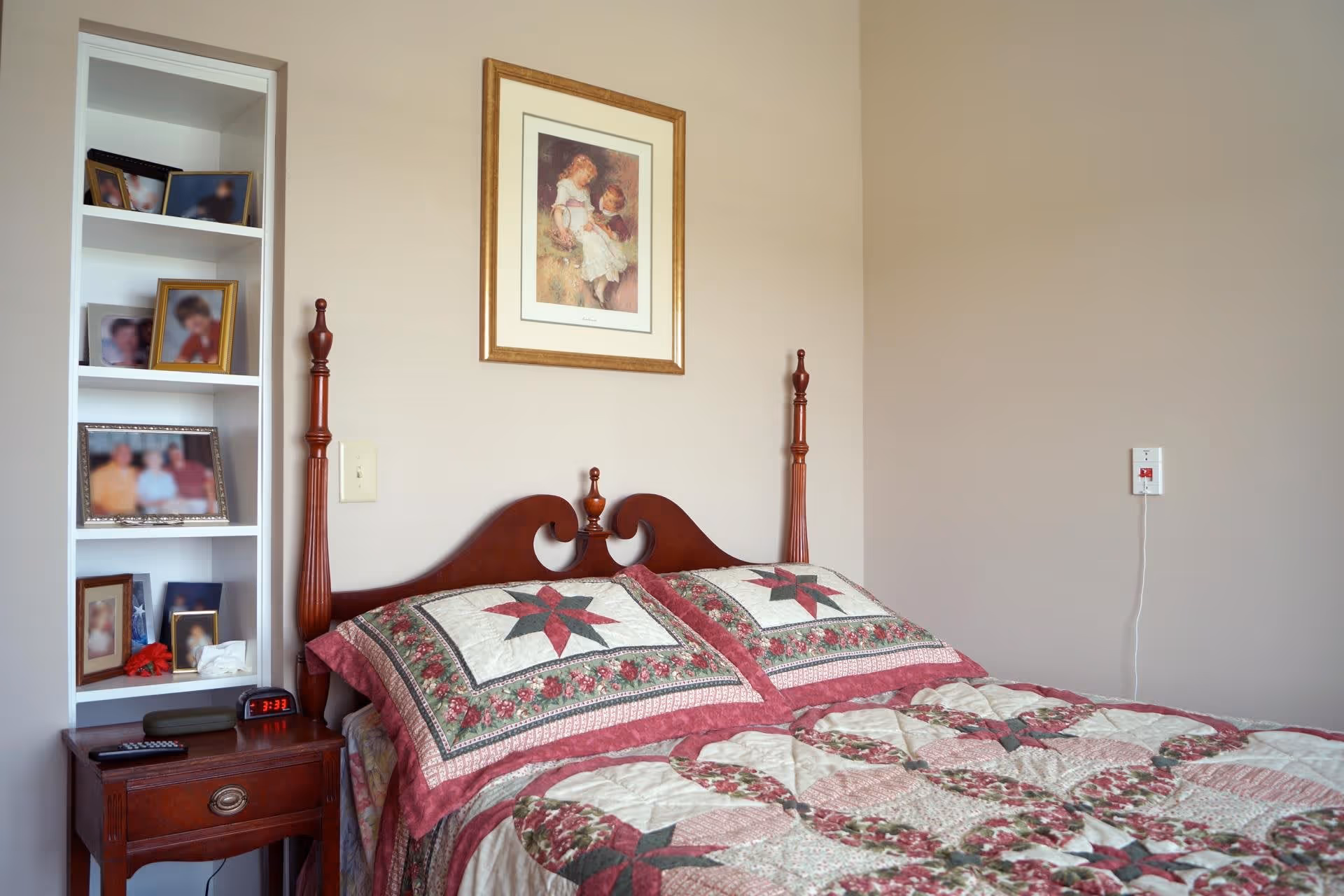 A bedroom with a wooden bed frame featuring two tall posts and a quilted bedspread with a floral and star pattern in shades of red, green, and cream. Above the bed hangs a framed picture of two children. To the left of the bed is a wooden nightstand with a digital alarm clock showing 3:33, a remote control, and a small shelf unit with several framed photos.