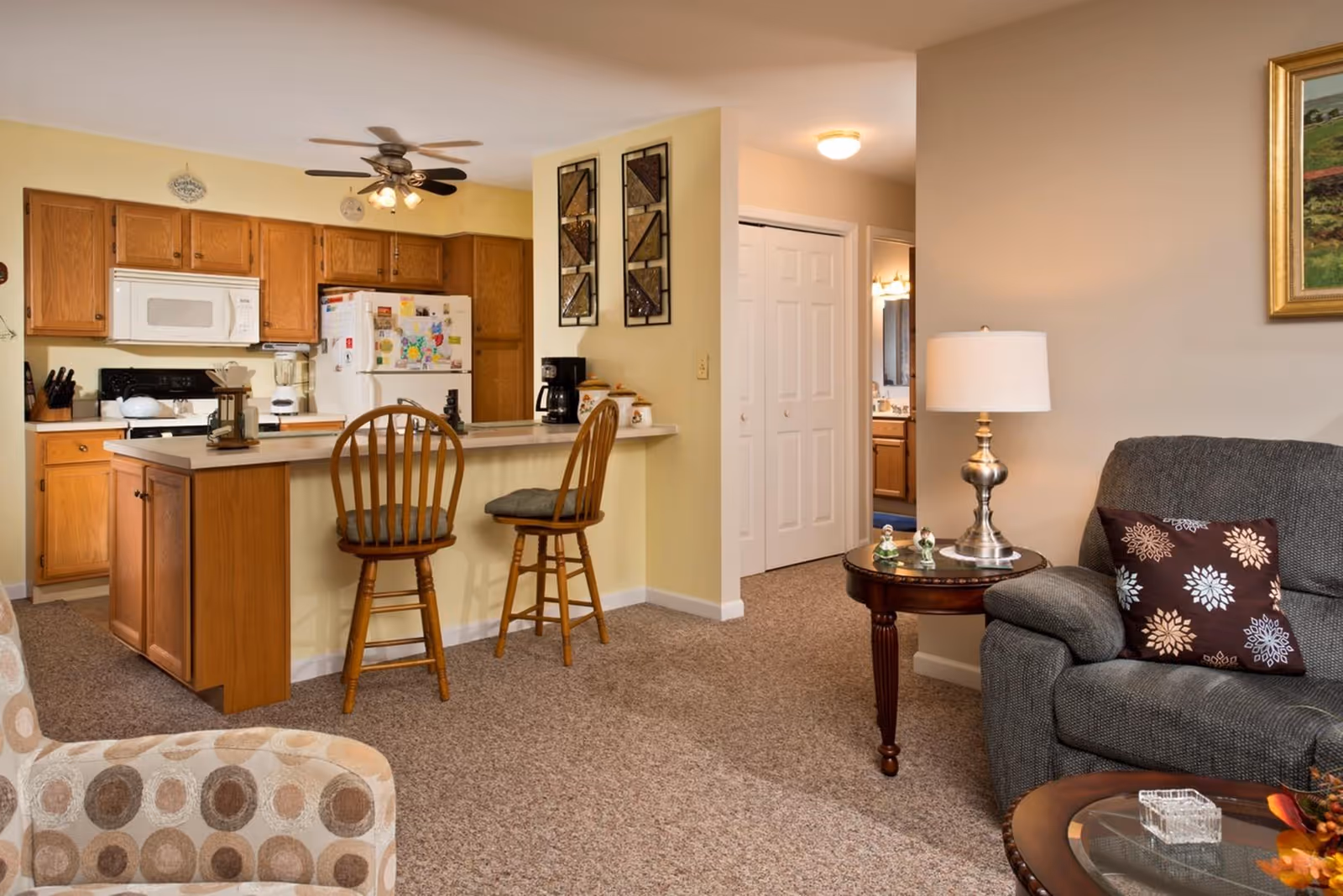 Interior view of a senior living facility showing a kitchen area with wooden cabinets, a white microwave, and a refrigerator covered with magnets and drawings. Two wooden bar stools are placed at the kitchen counter. Adjacent to the kitchen is a living area with a gray upholstered armchair, a round wooden side table with a lamp, and a patterned pillow. The carpeted floor and neutral wall colors create a warm and cozy atmosphere.