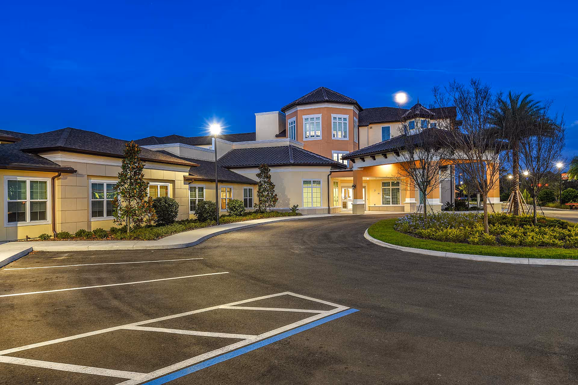 Front entrance of a senior living facility at dusk with a curved driveway, landscaped island, and parking spaces.