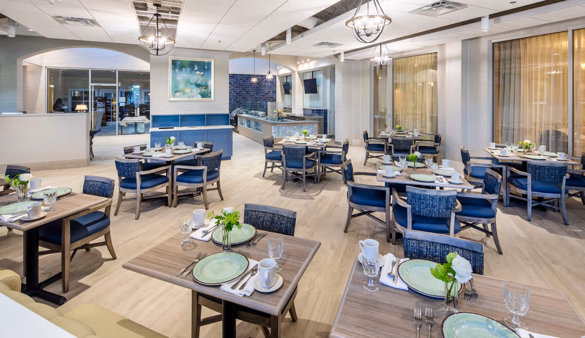 A bright and spacious dining room with multiple wooden tables set with green plates, white cups, glassware, and silverware. Each table has a small floral centerpiece. The room features light wood flooring, large windows with sheer curtains, and modern light fixtures hanging from the ceiling. In the background, there is a buffet area with a blue tiled wall and a framed artwork above a blue shelf.