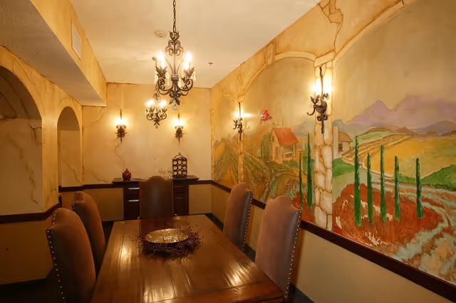 Dining room with a wooden table and upholstered chairs, ornate chandeliers, wall sconces, and a painted countryside mural on the walls.