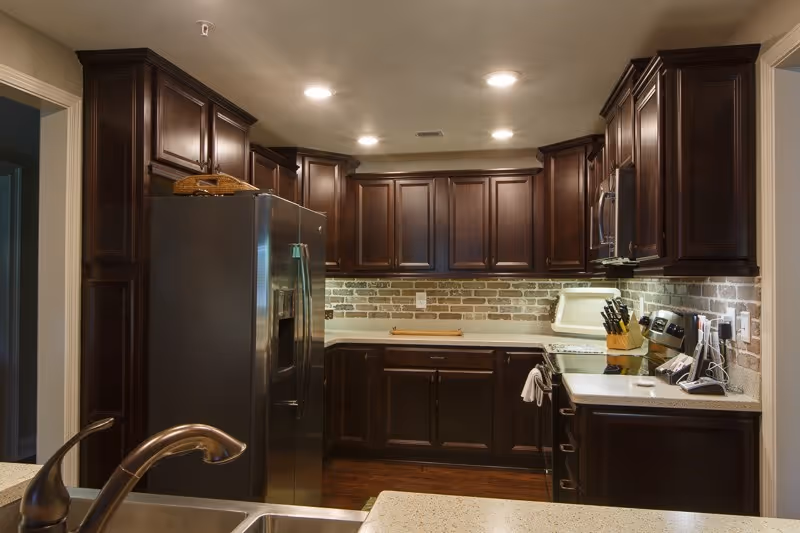 Modern kitchen with dark wood cabinets, stainless steel refrigerator, granite countertops and a sink in the foreground.