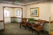 A quiet interior hallway area with two brown leather chairs and a small wooden table with a green plant on it. The walls are decorated with framed artwork and a floral wallpaper border near the ceiling. There is a door with a window and an exit sign above it at the end of the hallway.