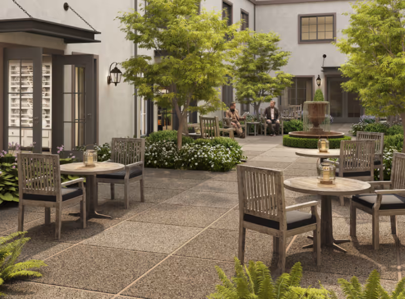Outdoor courtyard area with several wooden tables and chairs arranged on a paved surface. There are green trees and bushes surrounding the seating area, and a water fountain is visible in the background. Two people are sitting and conversing near the fountain. The building walls have windows and doors opening into the courtyard.
