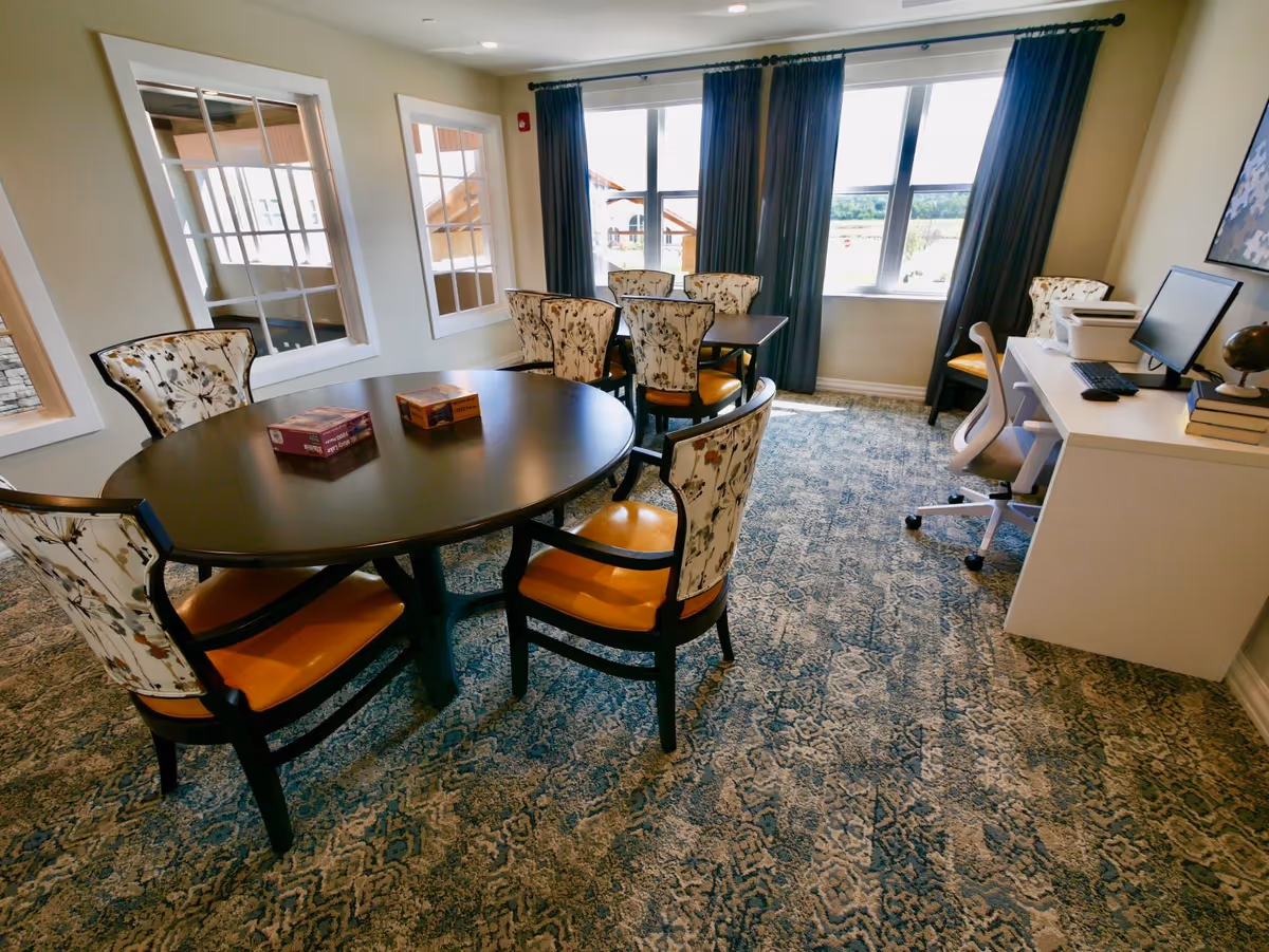 A well-lit room with two tables surrounded by floral-patterned chairs with orange seats. The room has large windows with dark curtains allowing natural light to enter. A desk with a computer, printer, and books is positioned against one wall. The floor is covered with a patterned carpet.