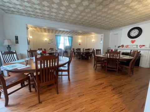 A spacious dining room with multiple wooden tables and chairs arranged neatly on a wooden floor. The room has a patterned ceiling, light-colored walls, a large clock above a white mantel, and windows with blue curtains letting in natural light. The tables have small decorative items and placemats.