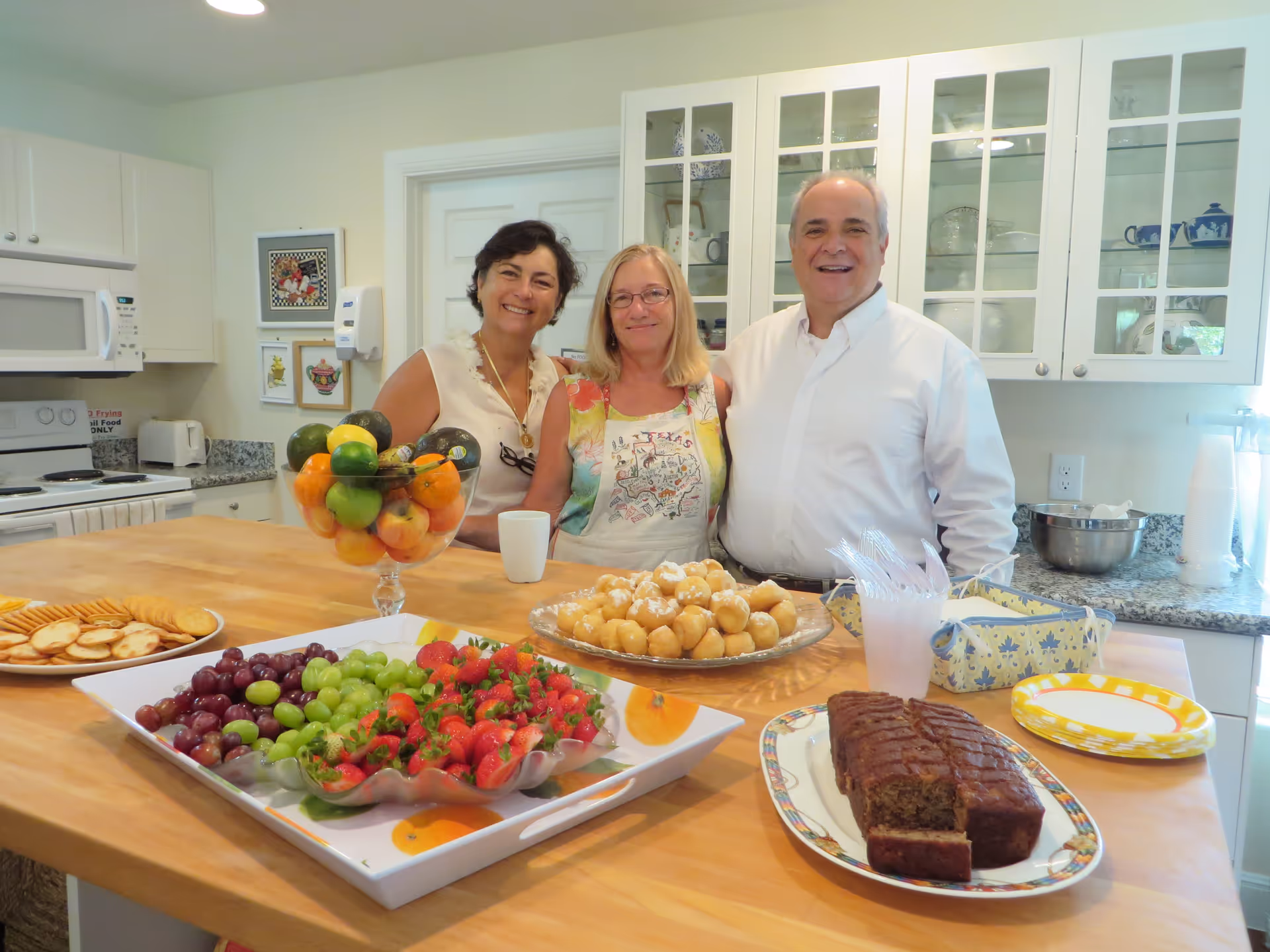 Three adults standing behind a kitchen island with a variety of food items including crackers, grapes, strawberries, a bowl of mixed fruit, cream puffs, and a loaf cake. The kitchen has white cabinets with glass doors and a microwave above the stove.