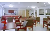 A bright and clean dining room with multiple wooden tables and cushioned chairs arranged neatly. Each table has a small red flower centerpiece. The room has white walls, large windows letting in natural light, and a counter area with cabinets in the background.