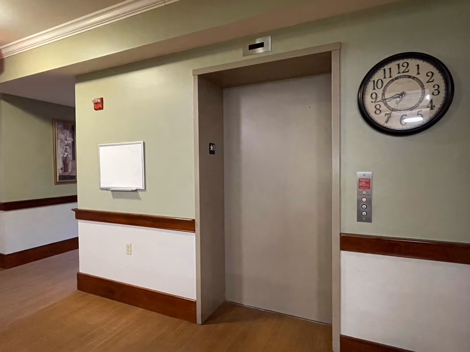 Interior hallway showing closed elevator doors flanked by a wall clock and a bulletin board.