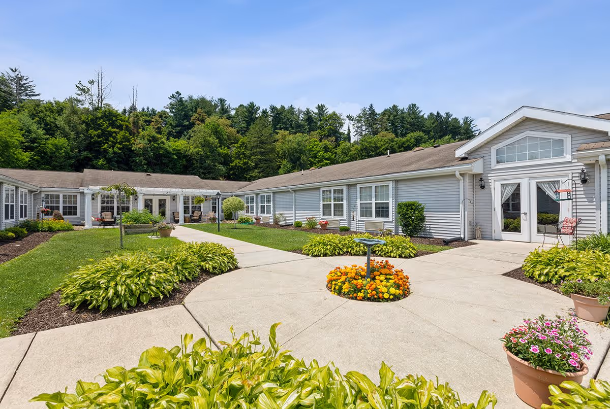 Outdoor courtyard area of a senior living facility with a circular concrete walkway surrounding a flower bed with orange and yellow flowers. The courtyard is bordered by green shrubs and potted plants, with a single-story building featuring large windows and glass doors in the background. Trees and a clear blue sky are visible behind the building.