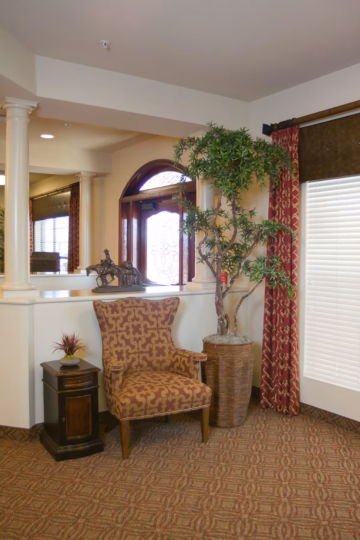 Decorative upholstered armchair beside a small wooden side table and a tall potted plant in a furnished entry/lounge area with curtains and columns.
