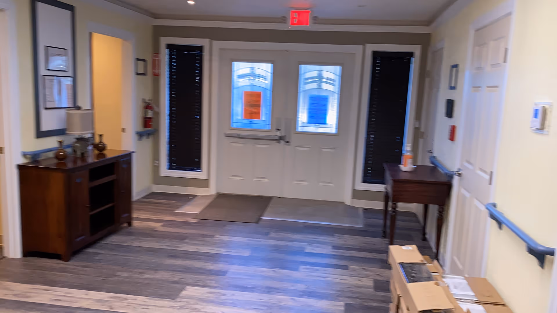 Interior view of a hallway in a senior living facility with double exit doors at the end, two windows with blinds on either side of the doors, wooden furniture including a cabinet and a small table, handrails along the walls, and a fire extinguisher mounted near an open doorway.