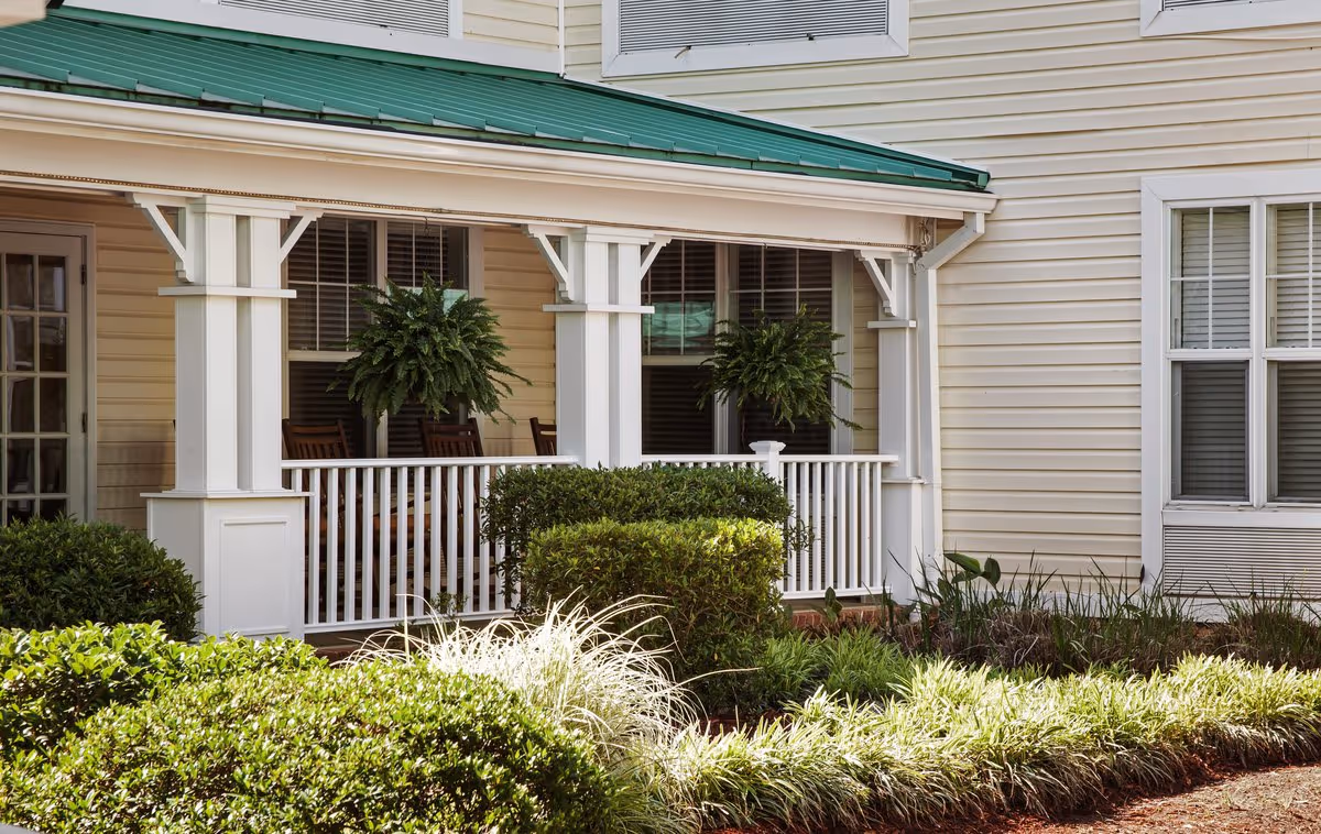 Front porch of a light-colored building with white railings, hanging ferns, rocking chairs, and manicured shrubs.