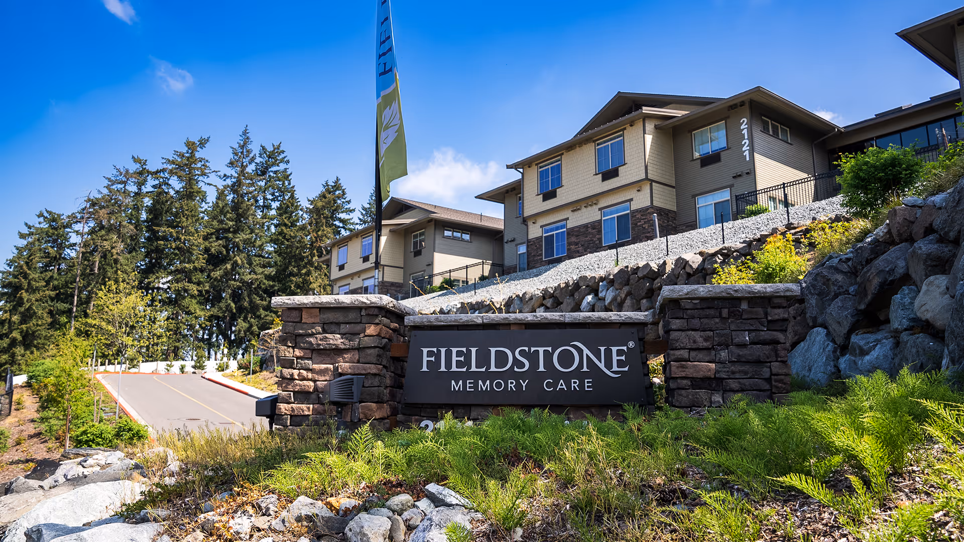 A stone sign reading "FIELDSTONE Memory Care" stands in front of a landscaped slope and a multi-story care building.