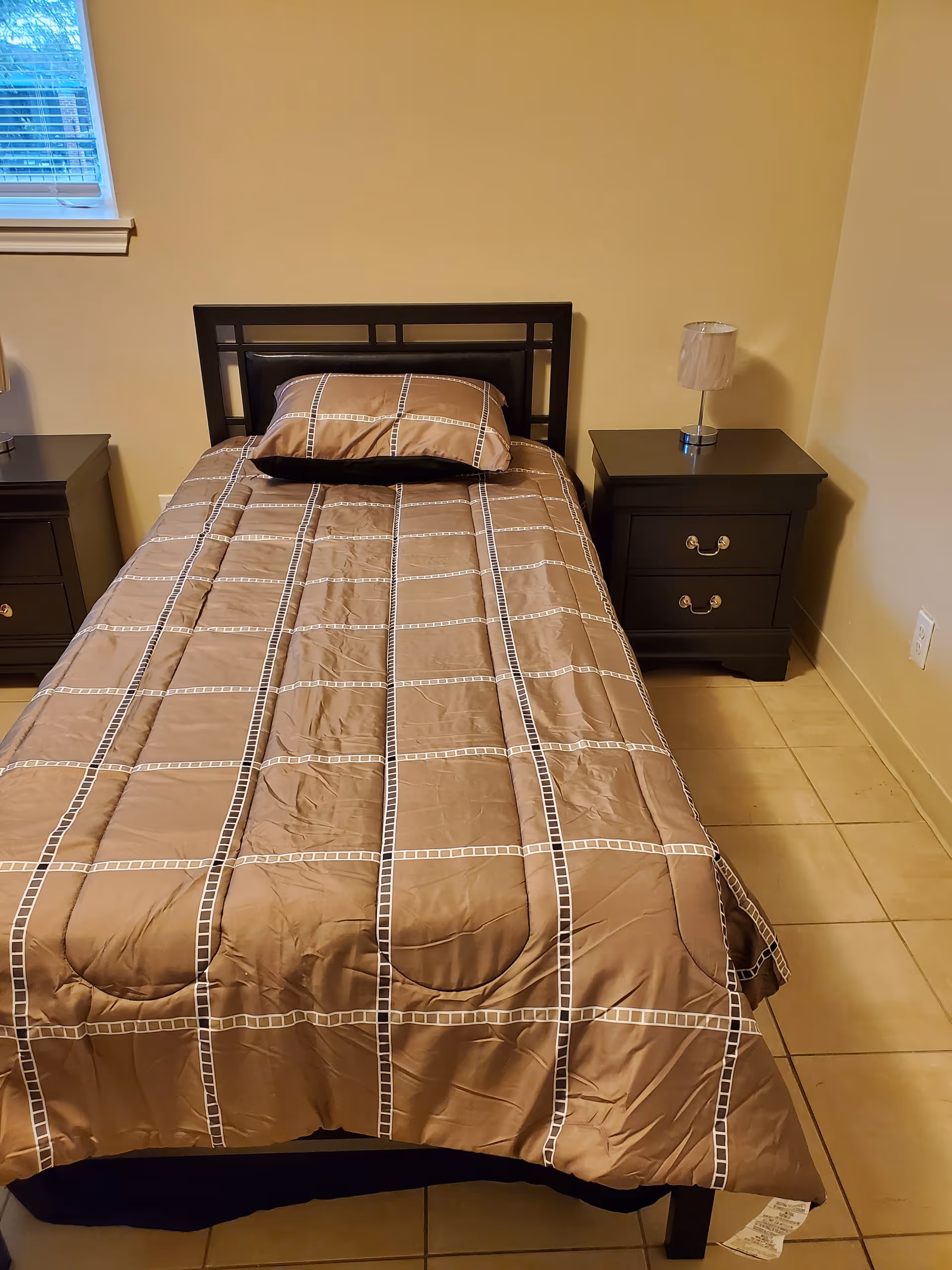 A single bed with a brown and beige checkered comforter and matching pillow is centered against a beige wall. On each side of the bed, there are dark wooden nightstands with drawers. The nightstand on the right has a small lamp with a white lampshade. A window with blinds is partially visible on the left side of the image.