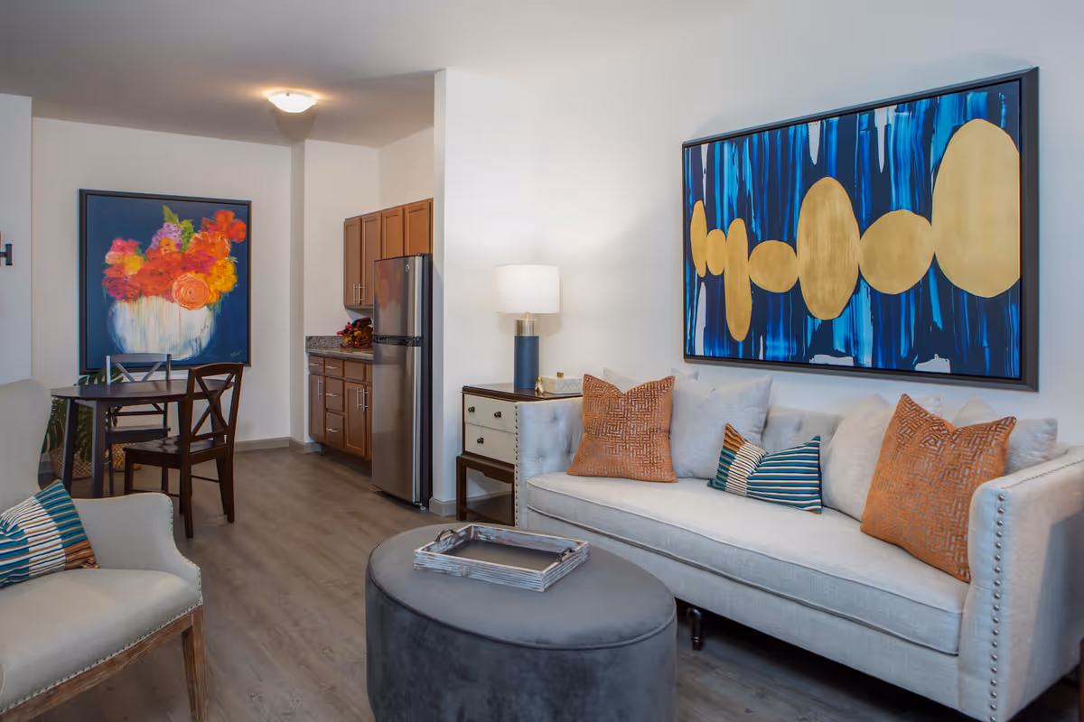 A modern living room area with a light gray sofa adorned with orange and striped pillows, a round gray ottoman with a wooden tray on top, and a beige armchair with a striped pillow. The wall behind the sofa features a large abstract painting with blue and gold colors. In the background, there is a small dining table with two chairs and a colorful floral painting on the wall. A kitchen area with wooden cabinets and a stainless steel refrigerator is visible further back.