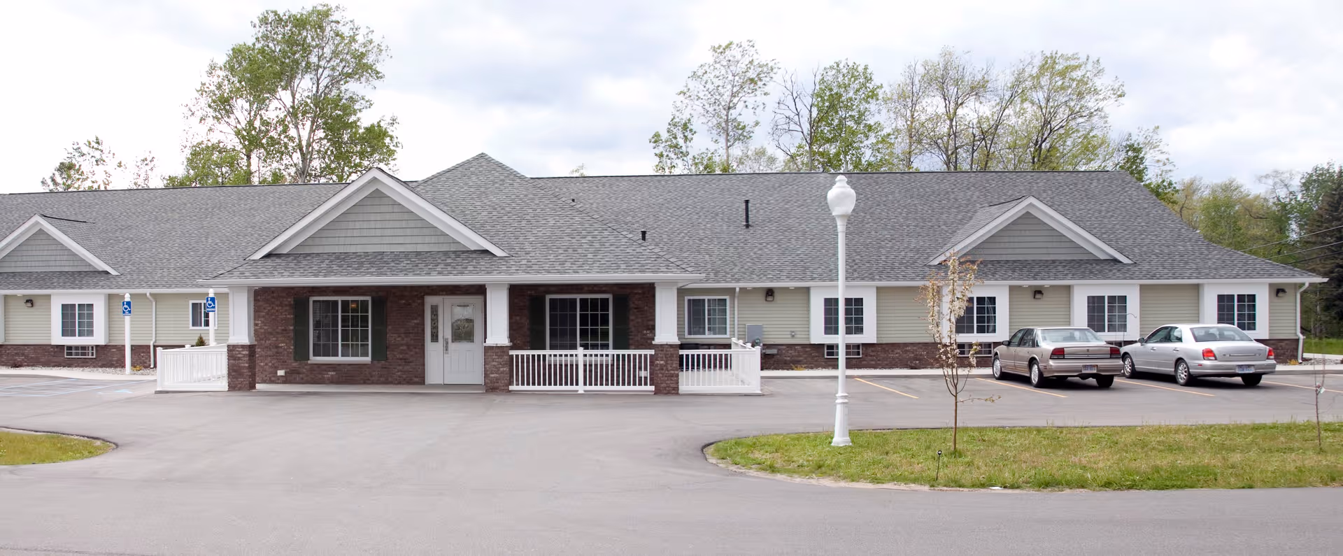 Exterior front view of a single-story senior living facility building with a gray roof, brick and light-colored siding, a white front door, windows, a small porch with white railings, a parking lot with two cars, and a street lamp in front. Trees and greenery are visible in the background under a cloudy sky.
