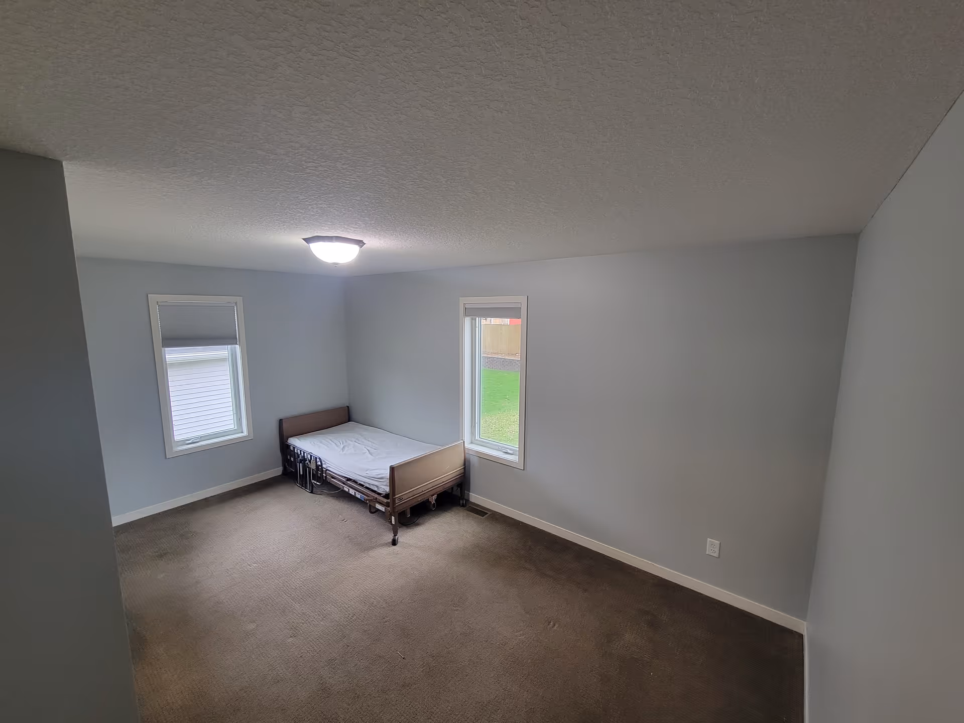 Sparsely furnished bedroom with a single medical-style bed against light gray walls, two windows, brown carpet, and a ceiling light.