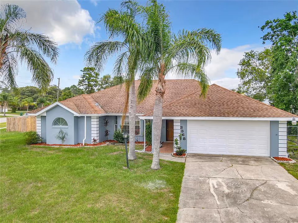 Single-story light-blue house with a brown roof, two tall palm trees in front, an attached garage and a driveway.