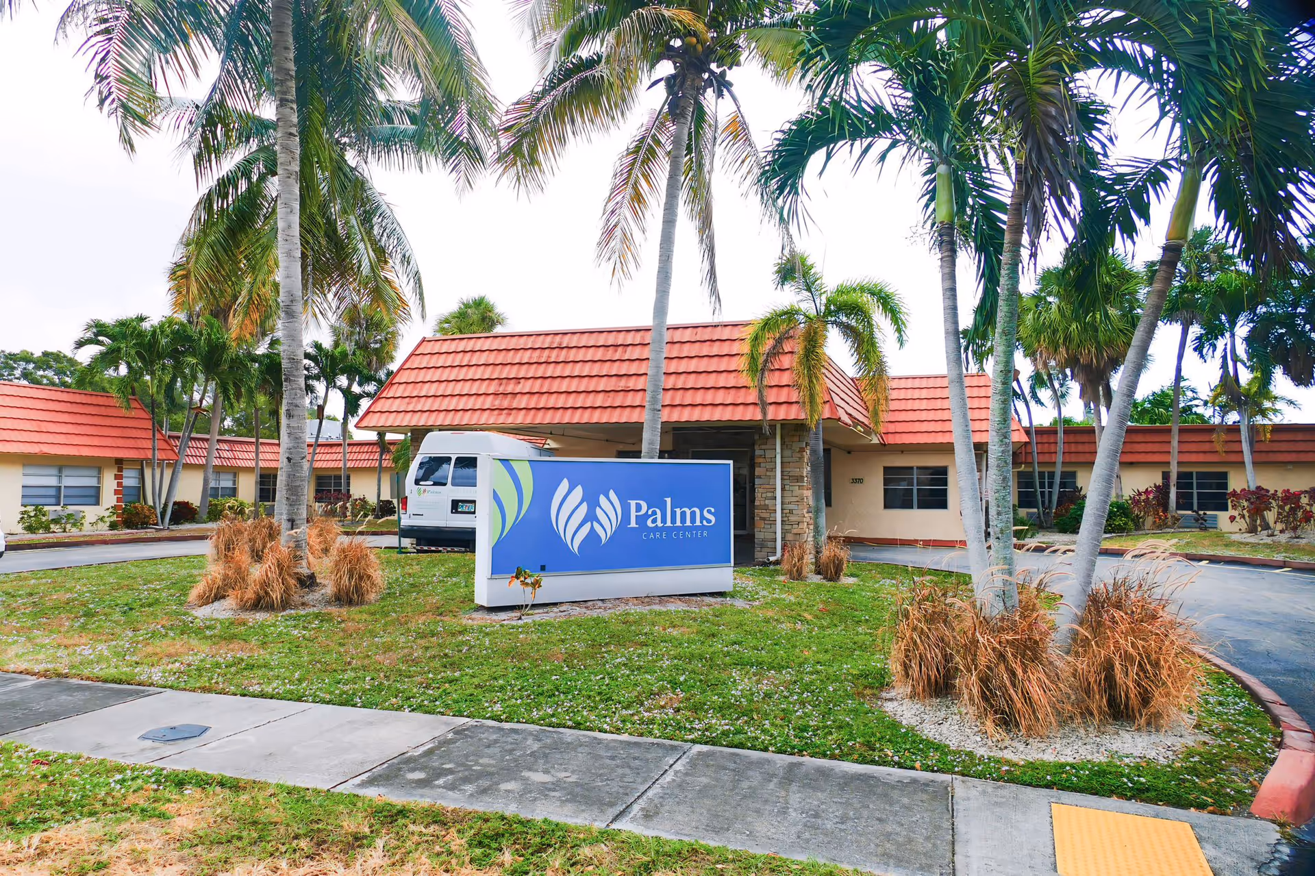 Exterior view of Palms Care Center with a red-tiled roof, palm trees, and a blue Palms Care Center sign in front.