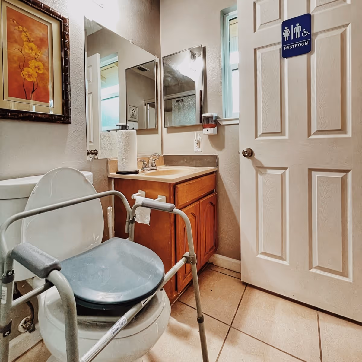A small restroom with a toilet equipped with a raised seat and armrests for accessibility, a wooden vanity with a sink, a paper towel roll on the counter, a wall-mounted soap dispenser, a mirror above the sink, a window, and a door with a restroom sign indicating accessibility.