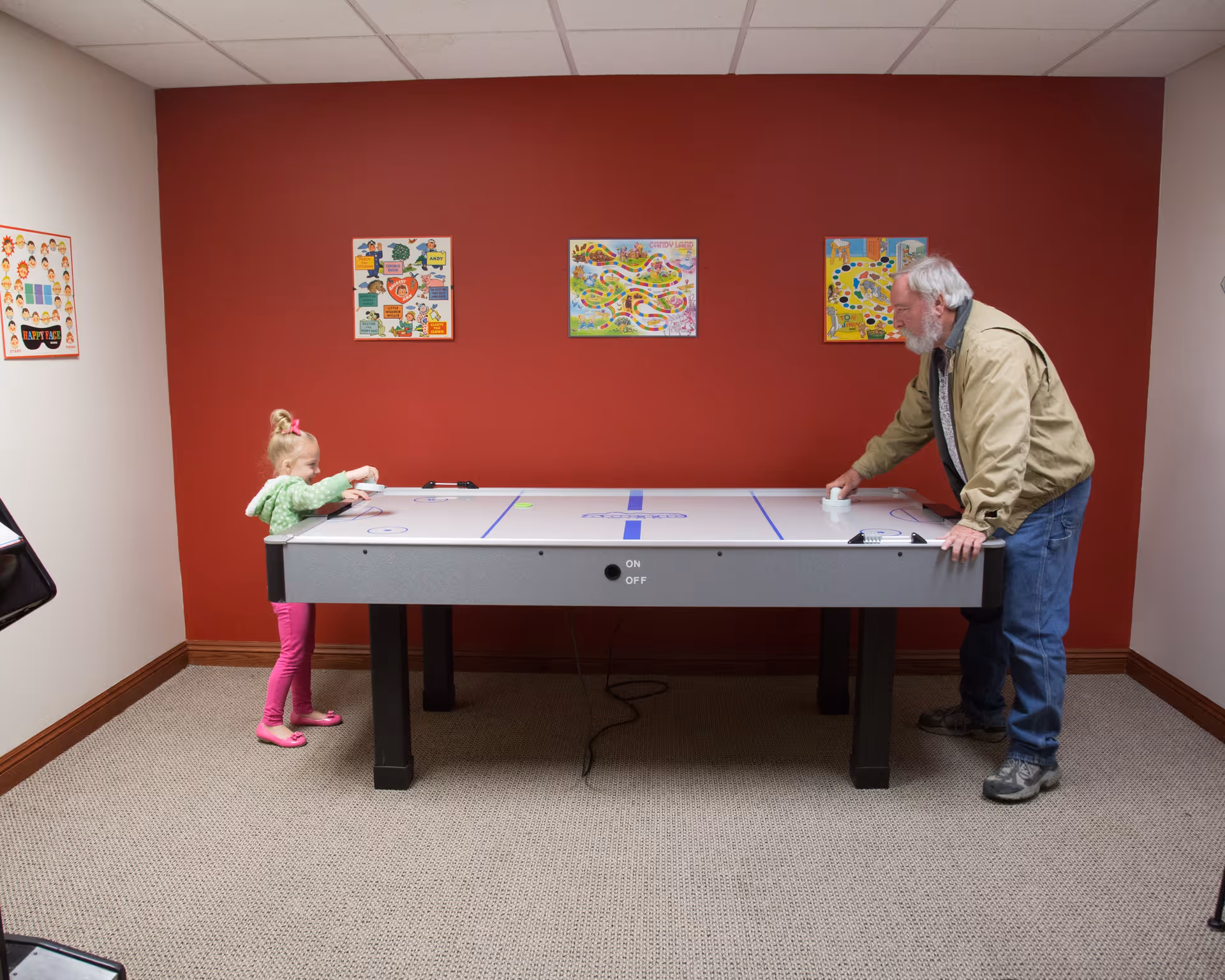 An elderly man and a young girl playing air hockey in a room with a red accent wall and three colorful posters. The floor is carpeted and the room has a drop ceiling with fluorescent lighting.