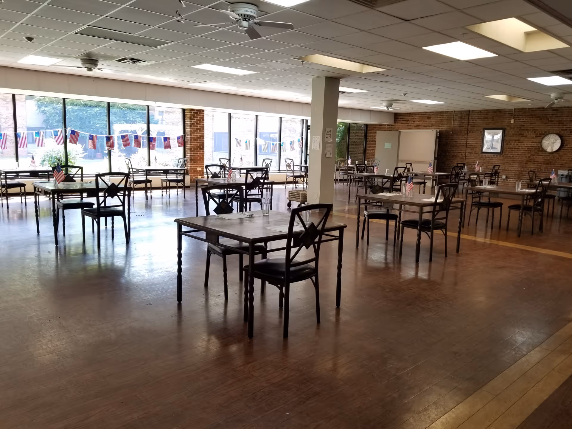 A spacious dining room with multiple tables and chairs arranged neatly. Each table has a small American flag and some have glasses and utensils. Large windows along one wall let in natural light, and there are American flag decorations hanging across the windows. The room has a wooden floor, brick walls, ceiling fans, and fluorescent lighting.