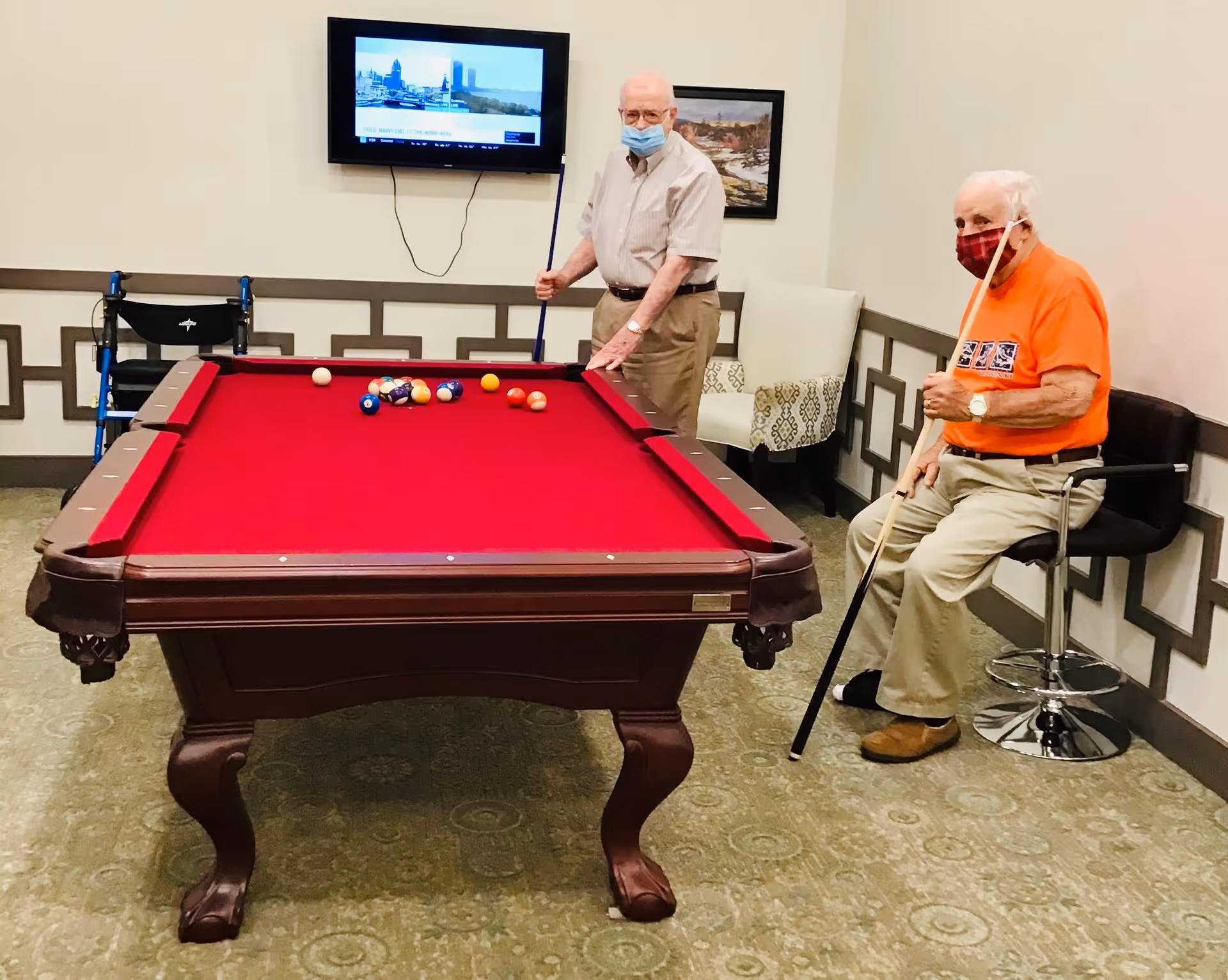 Two elderly men wearing face masks playing pool in a room with a red pool table. One man is standing and holding a pool cue, while the other is sitting on a stool holding a pool cue. A walker is visible near the standing man. A TV is mounted on the wall behind them, and a framed picture hangs next to the TV.
