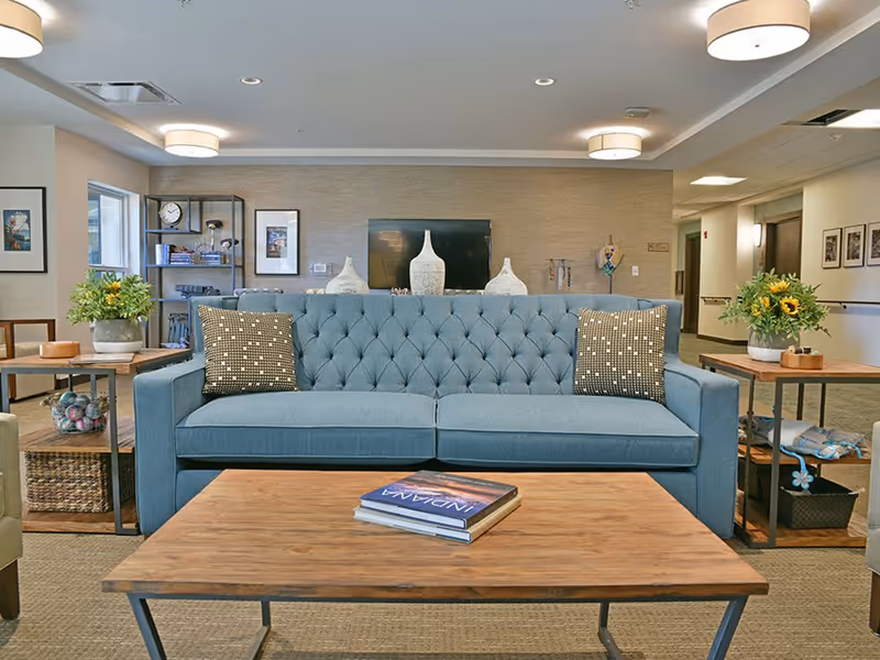 Communal living room with a blue tufted sofa, wooden coffee table, side tables with plants, and a TV on the back wall.