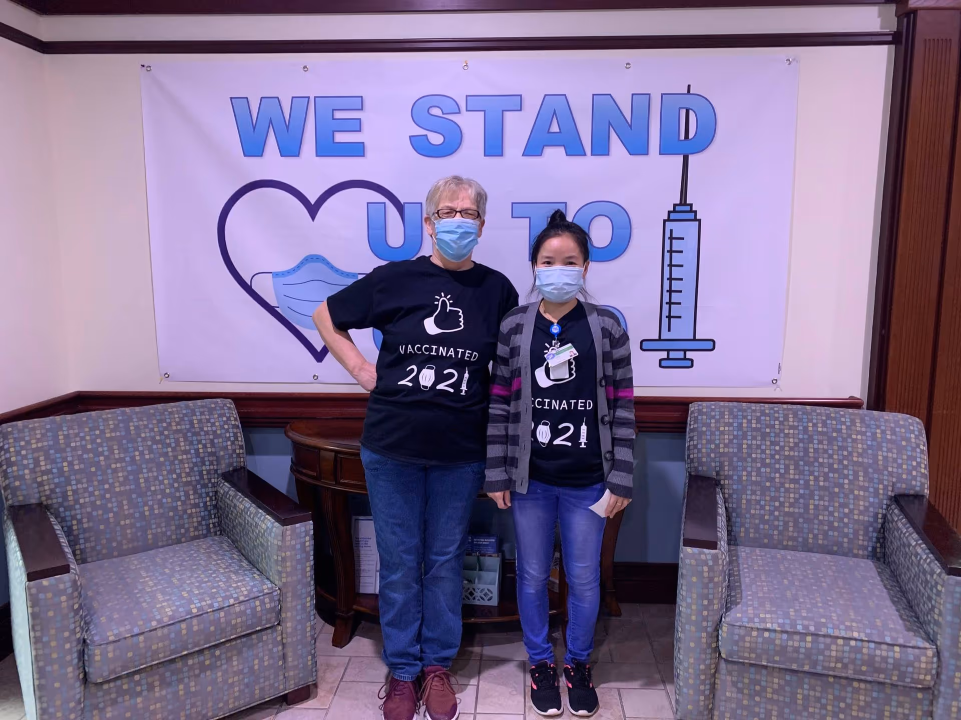 Two people wearing face masks and black t-shirts that say 'Vaccinated 2021' standing between two patterned armchairs in a room. Behind them is a large banner with a heart, a face mask, a syringe, and the words 'WE STAND UP TO'.