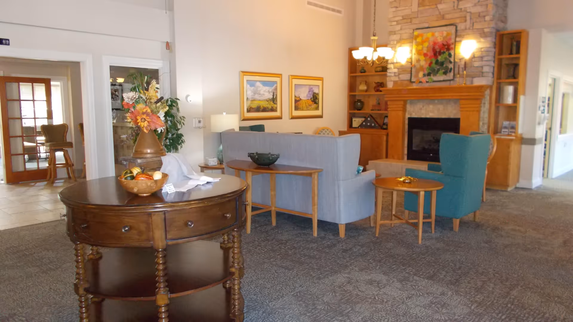 A cozy living room area with a gray sofa, teal armchairs, a wooden round table with a bowl of decorative gourds, and a stone fireplace with a colorful painting above it. The room has carpeted flooring, framed landscape paintings on the wall, and built-in wooden shelves with decorative items.