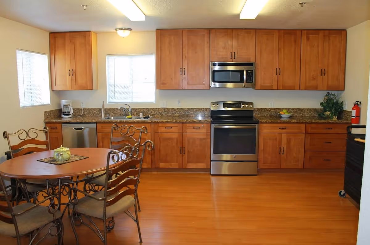 A kitchen area with wooden cabinets, a stainless steel oven and microwave, a dishwasher, a coffee maker, and a round wooden dining table with four chairs. The room has two windows with blinds and a fire extinguisher on the right side.