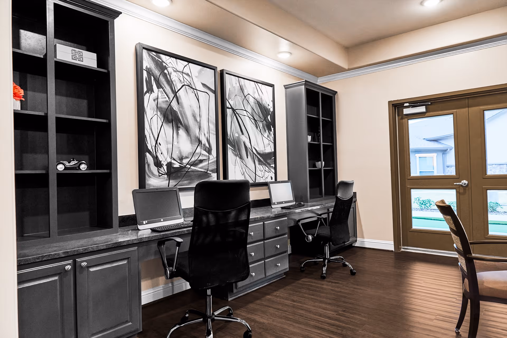 Interior view of a workspace area with two black office chairs in front of a long desk with two computers. The desk has built-in cabinets and shelves, some with decorative items. Two abstract black and white paintings hang on the wall above the desk. There is a door with windows leading outside, and a wooden chair is partially visible on the right side.