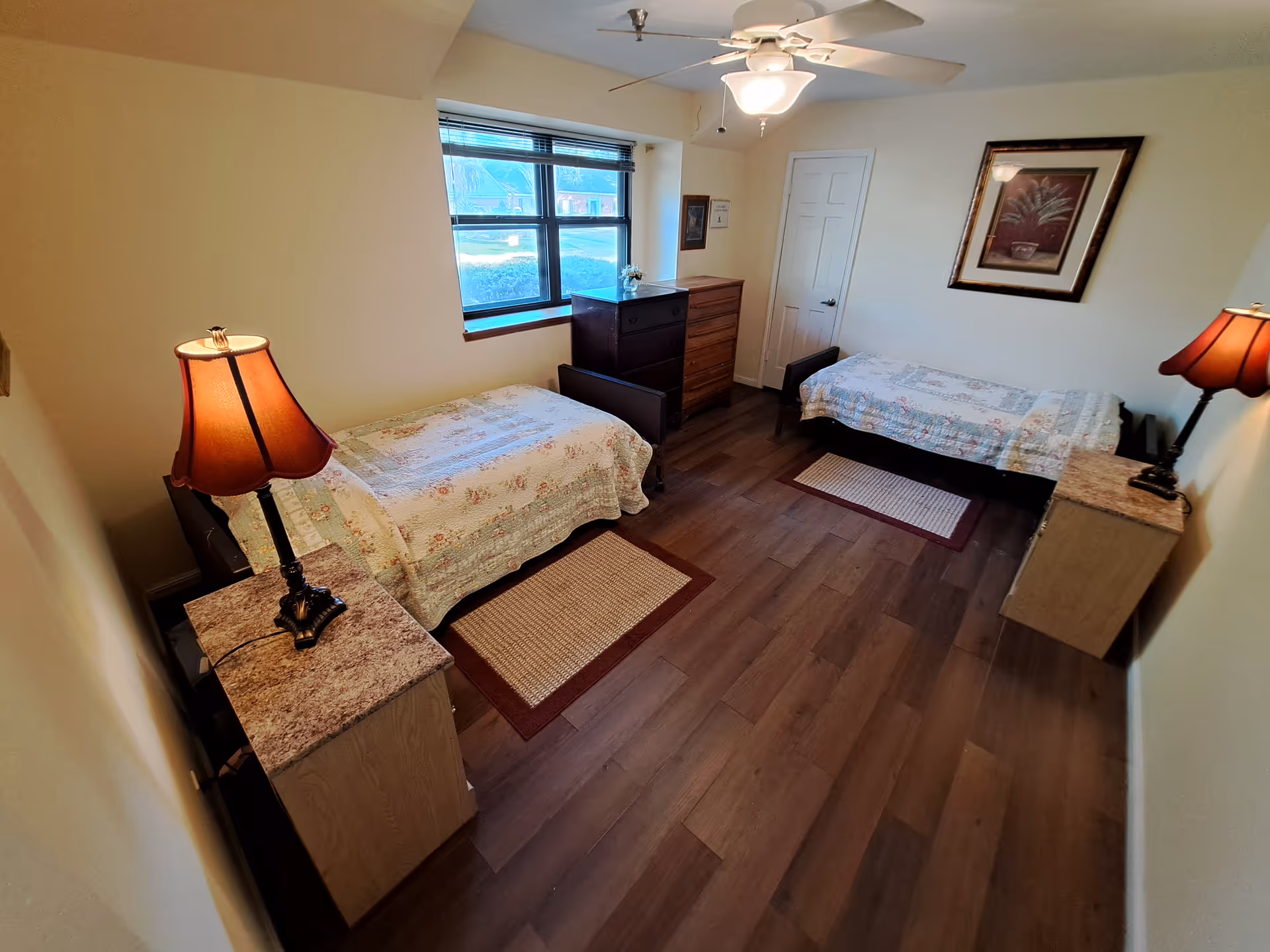 Bright twin bedroom with two single beds, nightstands and lamps, a dresser under a window, ceiling fan, and framed artwork.