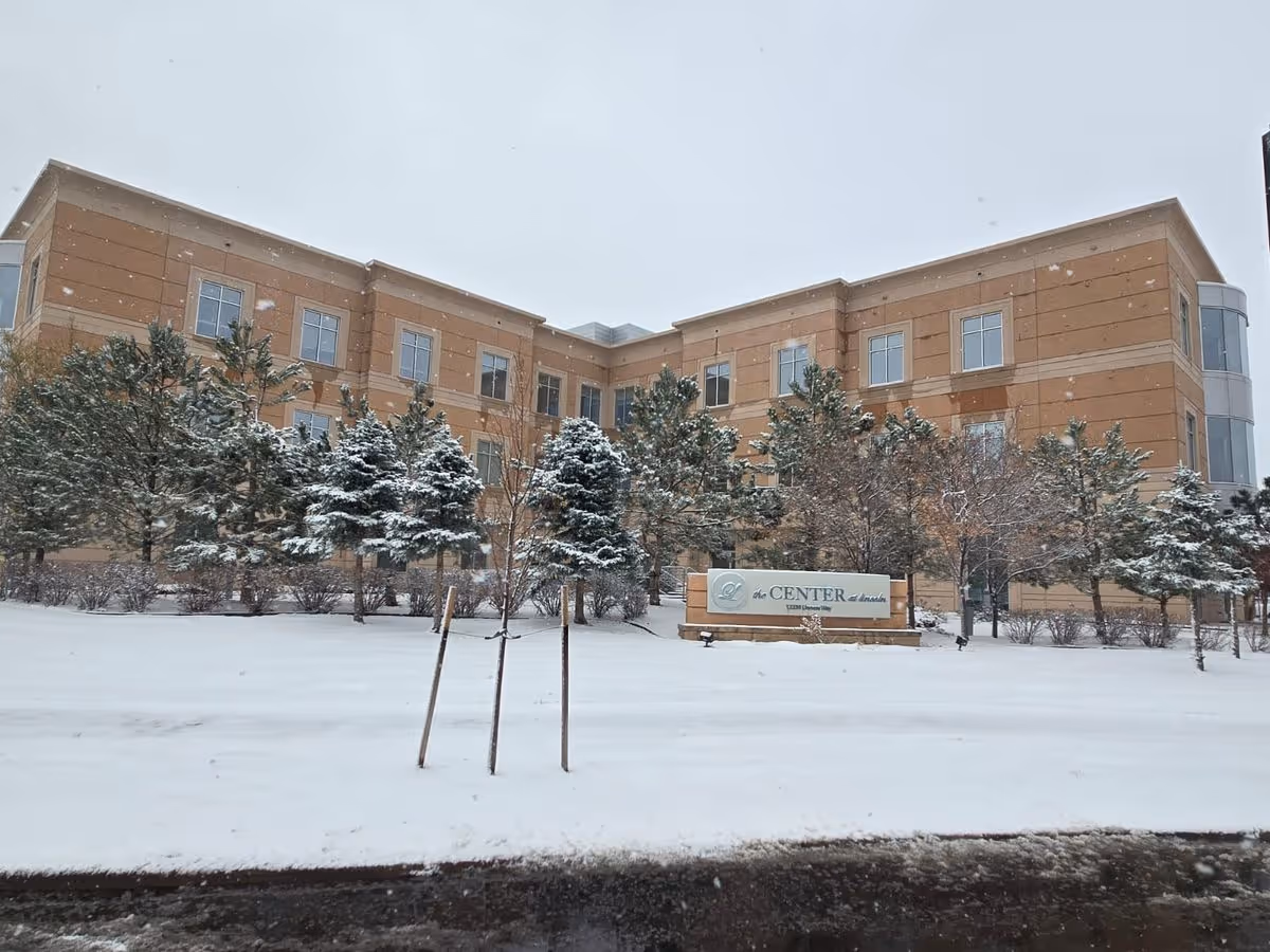 Snow-covered front exterior of a multi-story senior living building with trees and a sign reading "Center".