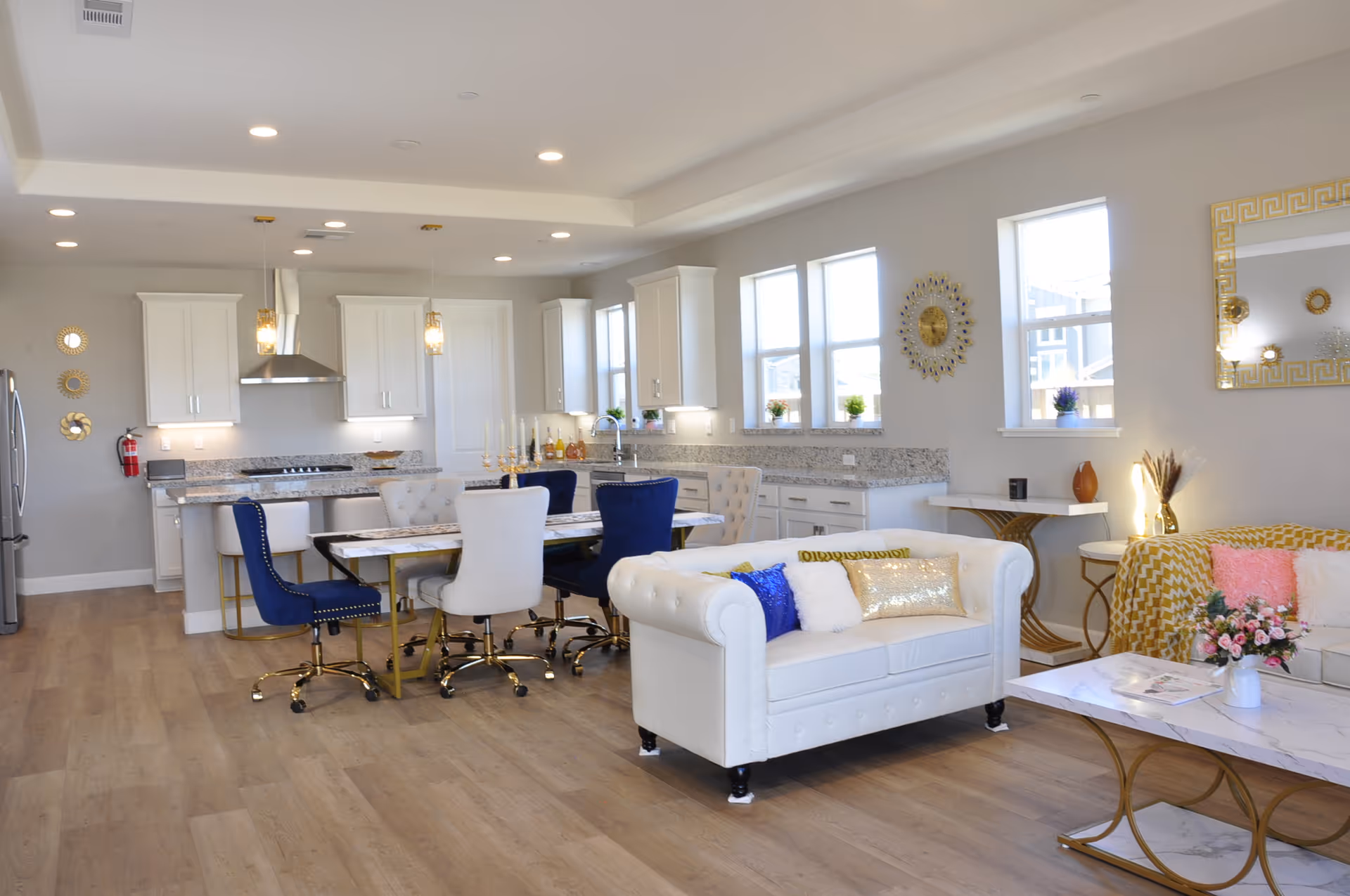 Bright open-concept living room and kitchen with white sofas, blue and gold-accented dining chairs, and a marble-top coffee table.