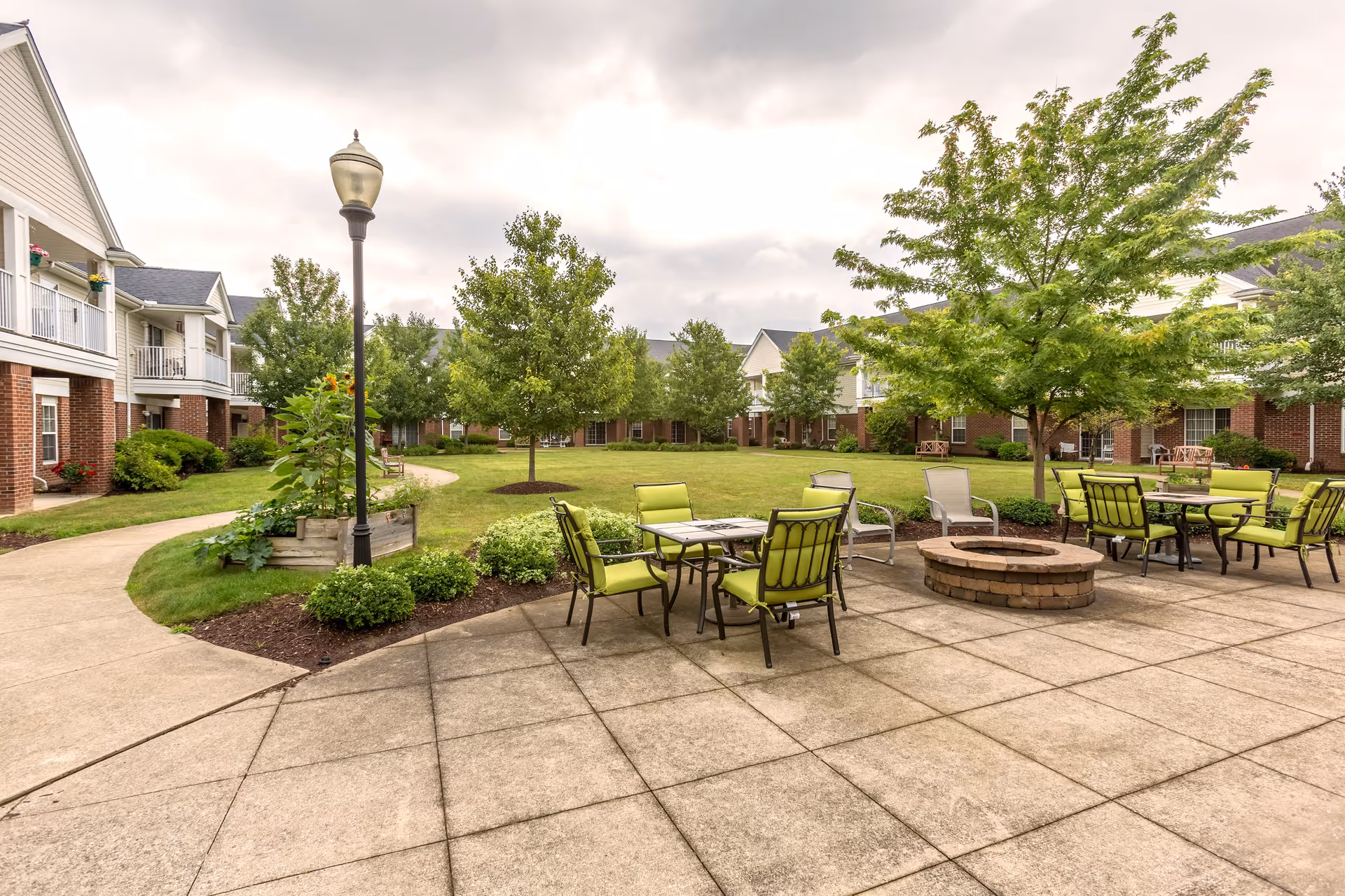 Outdoor courtyard area at Independence Village of Aurora featuring a paved patio with green cushioned chairs and tables arranged around a circular fire pit. The courtyard is surrounded by well-maintained grass, trees, and shrubs, with two-story residential buildings in the background under a cloudy sky.