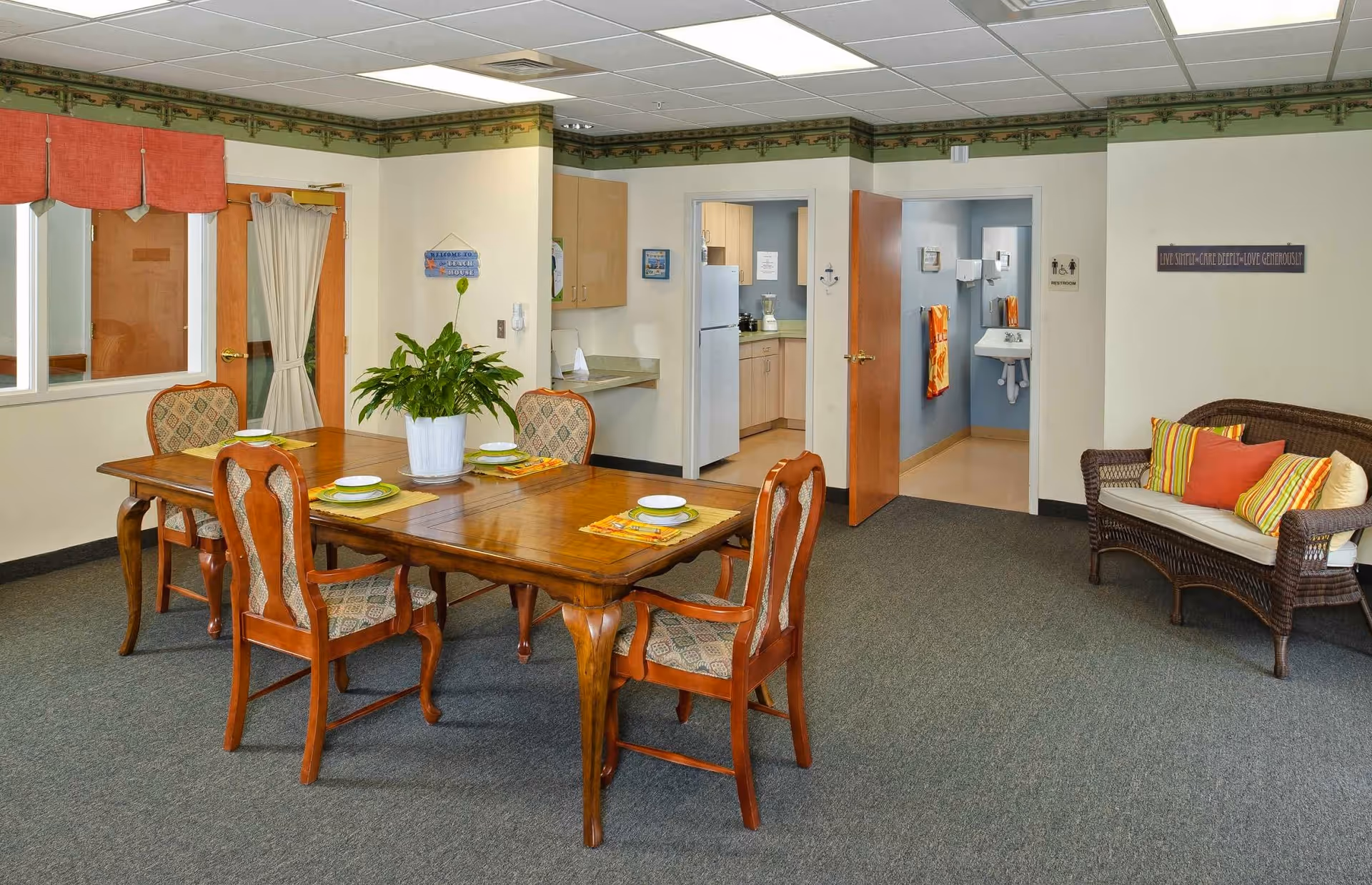 A cozy dining area with a wooden table set for four, featuring patterned chairs and a potted plant centerpiece. To the right, there is a wicker loveseat with colorful striped and solid pillows. In the background, an open door reveals a small kitchen with light wood cabinets and a refrigerator, and another open door leads to a restroom with a sink and hand towels. The room has a carpeted floor and cream-colored walls with a decorative border near the ceiling.