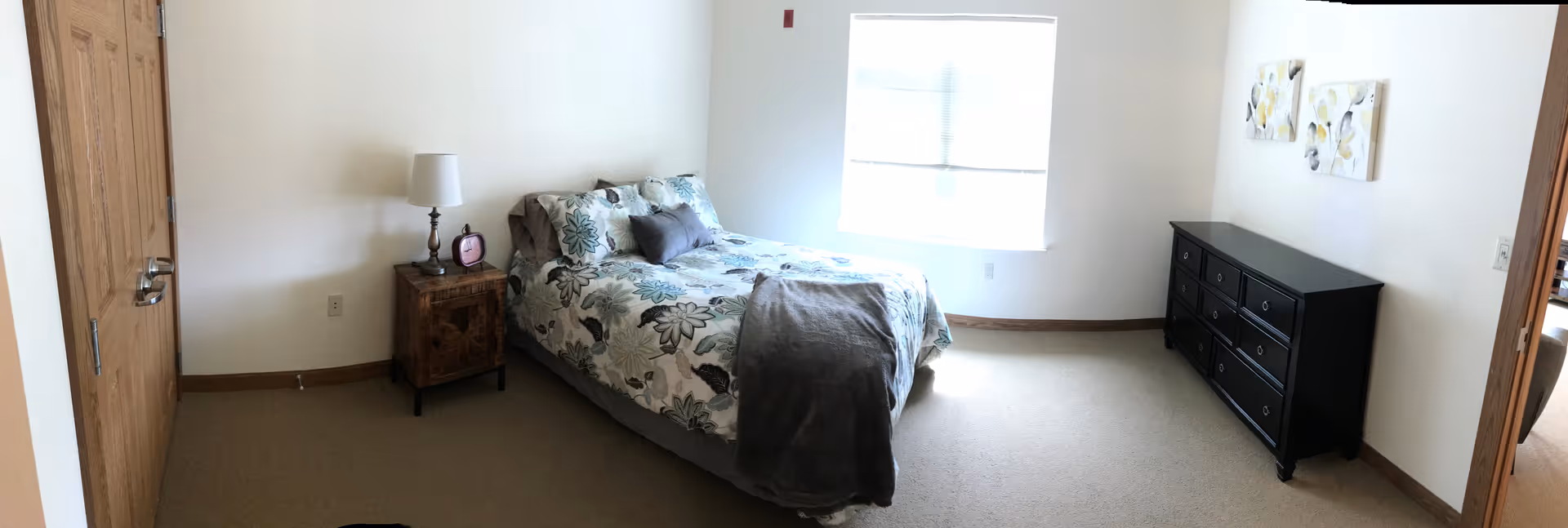 A bedroom with a bed covered in floral bedding and a gray throw blanket. There is a wooden nightstand with a lamp and a small clock next to the bed. A large window lets in natural light. Across from the bed is a black dresser with multiple drawers and two floral paintings hanging above it. The room has beige carpet and white walls with wooden trim.