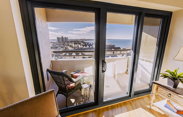 View through large sliding glass doors onto a balcony with a wicker chair and small table. The balcony overlooks a waterfront area with buildings and a body of water under a partly cloudy sky. Inside, part of a wooden side table with a plant and books is visible along with a chair.