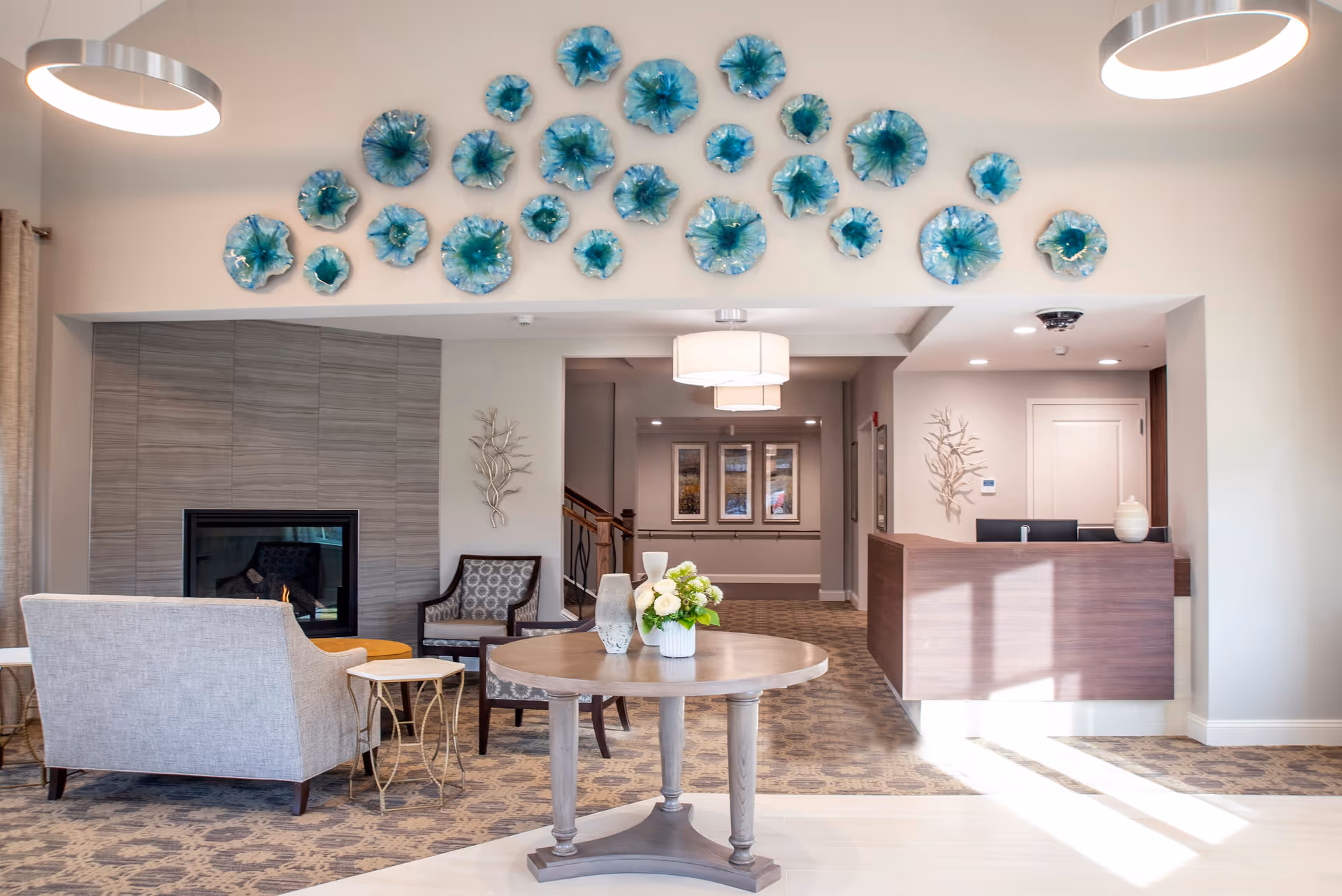 A bright and welcoming senior living facility lobby with a round wooden table in the center holding decorative vases and a flower arrangement. Behind the table is a seating area with a gray armchair, a patterned chair, and a modern fireplace set in a tiled wall. The back wall features a reception desk with two computer monitors and decorative wall art. Above the seating area, there is a wall installation of blue and green glass flowers. The space is well-lit with modern ceiling lights and natural sunlight streaming in.