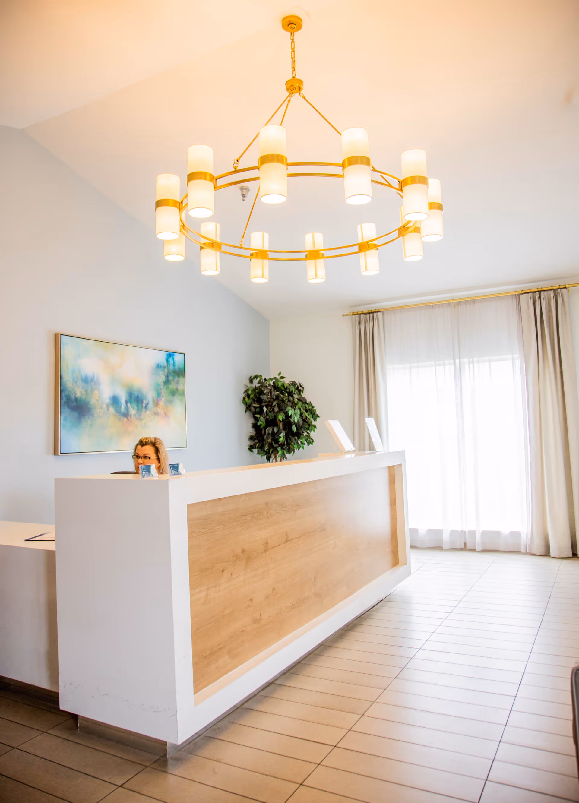 Reception area with a modern white and wood front desk, a woman sitting behind the desk, a large circular chandelier with multiple lights hanging from the ceiling, a potted plant, a colorful abstract painting on the wall, and large windows with sheer curtains allowing natural light to fill the space.
