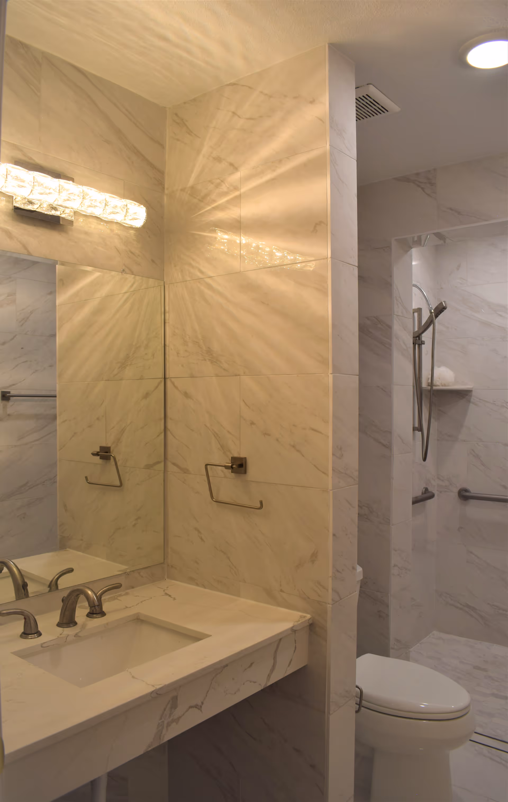 A modern bathroom with marble-patterned walls and countertop. The bathroom features a sink with a silver faucet, a large mirror above the sink, a toilet, and a walk-in shower with a handheld showerhead and grab bars. The lighting fixture above the mirror emits a warm glow.