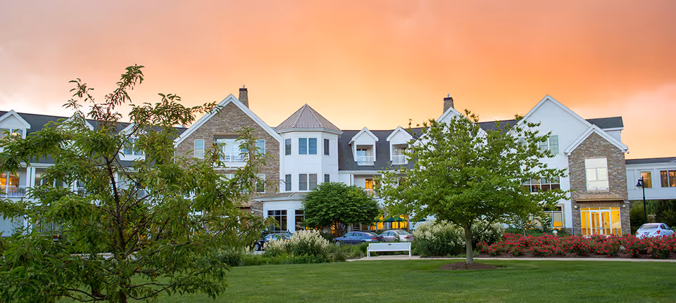 Exterior view of South Franklin Circle, a Judson Senior Living Community, showing a large multi-story building with stone and white siding, surrounded by green trees, bushes, and a well-maintained lawn under a colorful sunset sky.