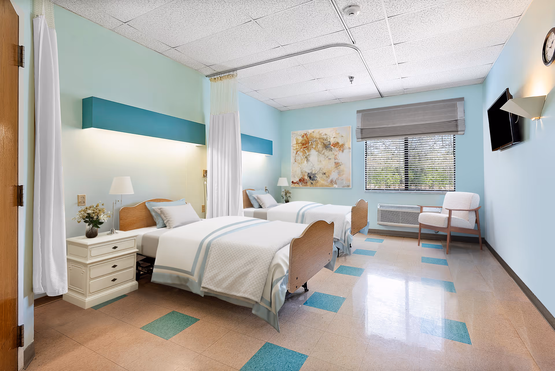 A bright and clean shared bedroom in a senior living facility with two single beds separated by a white curtain. Each bed has white bedding with light blue accents and a wooden headboard. There is a nightstand with a lamp and a vase of flowers between the beds. The room has light blue walls, a large window with blinds, a wall-mounted TV, a chair, and a colorful abstract painting on the wall.