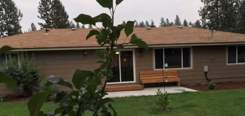 Single-story home's exterior showing a sliding glass door, a wooden bench on a small patio, and a grassy yard with plants in the foreground.