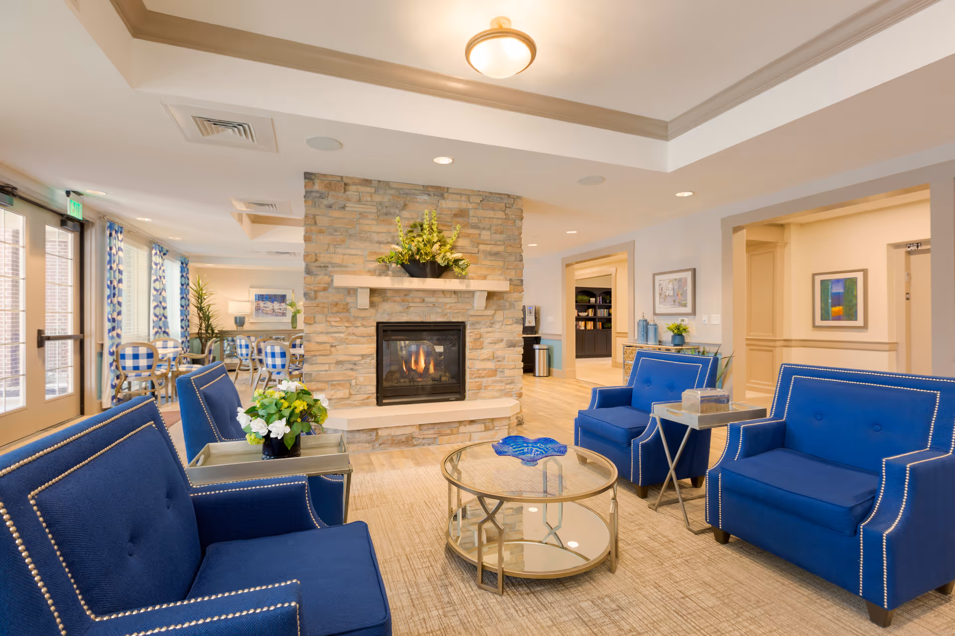 A cozy senior living facility common area featuring a stone fireplace with a fire burning, surrounded by four blue upholstered armchairs with nailhead trim. A round glass coffee table with a blue decorative piece sits in the center. In the background, there are dining tables with blue and white checkered chairs near large windows with patterned curtains, and a hallway leading to other rooms.