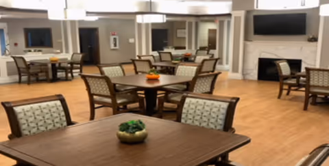 A spacious and well-lit common area with multiple wooden tables and cushioned chairs arranged neatly. The room features a fireplace with a large flat-screen TV mounted above it, light-colored walls, and hardwood flooring. Small decorative plants are placed on the tables, and there are large windows and doorways visible in the background.