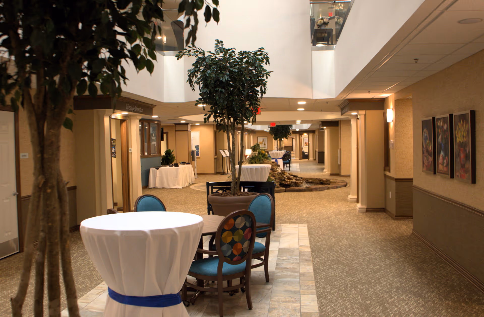 Interior hallway of a retirement community with beige walls and carpeted floors. There are several round tables covered with white tablecloths and blue ribbons, surrounded by chairs with colorful patterned backs. Large potted plants are placed along the hallway, and framed artwork hangs on the walls. The ceiling has recessed lighting and a skylight above.