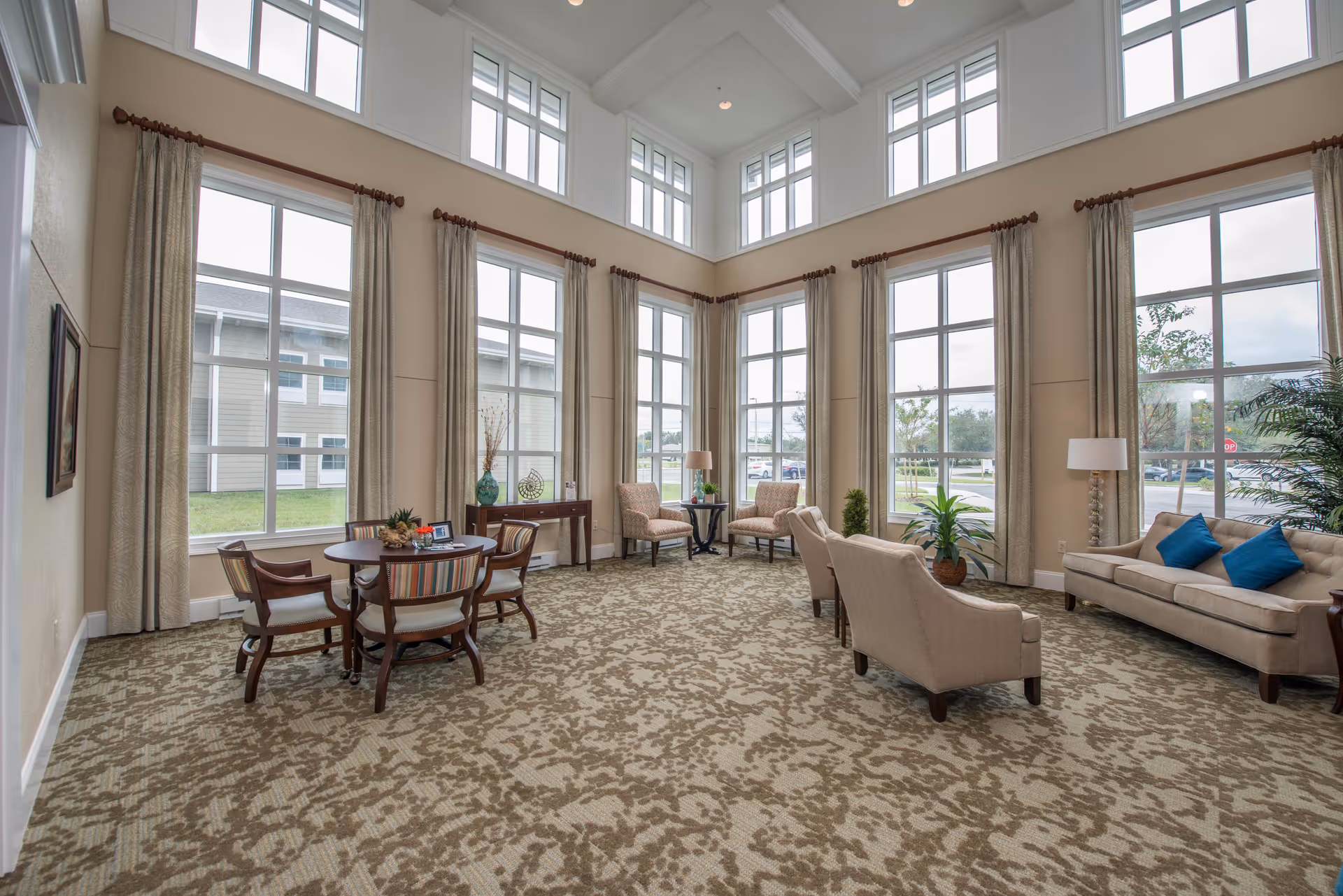 A spacious and well-lit living room area with large windows extending to the ceiling, beige patterned carpet, beige walls, and light-colored curtains. The room is furnished with a beige sofa with blue cushions, two beige armchairs, a round table with four chairs, a small side table with a lamp, and decorative plants.